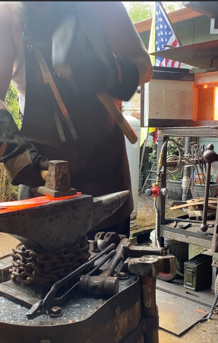 Tulos striking a glowing steel billet on an anvil beside the forge